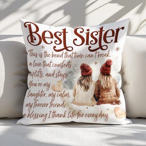 May include: White square throw pillow with the words "Best Sister" in brown script. The pillow features an illustration of two figures in winter attire, with text about sisterhood. The pillow is on a white surface.