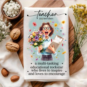 May include: A white tea towel with a colorful illustration of a teacher holding a bouquet of flowers and a clipboard. The text "Teacher (n) tee-cher: a multi-tasking educational rockstar who lives to inspire and loves to encourage" is printed on the towel.