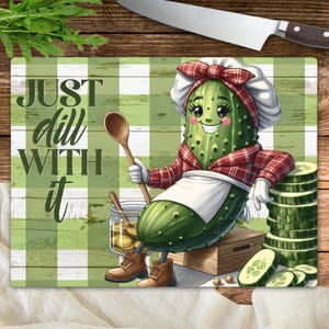 May include: A green pickle wearing a red and white checkered dress and a white apron, holding a wooden spoon. The pickle is sitting on a wooden box with a green and white checkered background. The text "Just dill with it" is written in black on the background.