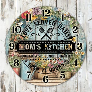 Könnte beinhalten: Eine runde Holzuhr mit floralem Design. Das Zifferblatt der Uhr hat einen blauen Hintergrund mit dem Text "Love Served Daily" und "Mom's Kitchen" in einem schwarzen Banner. Die Uhrzeiger sind schwarz und die Zahlen sind schwarz und weiß.