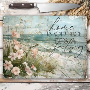 Könnte beinhalten: Rechteckiges Schneidebrett mit einer Küstenlandschaft mit rosa Blumen, Strandgras und Meereswellen. Der Text "home is not a place... it's a feeling" ist in schwarzer Schrift geschrieben. Ein Messer mit braunem Griff liegt obenauf.