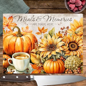 May include: A decorative cutting board with a fall theme. It features a large orange pumpkin, sunflowers, and the text "Meals & Memories ARE MADE HERE." A white coffee cup and a knife are also visible.