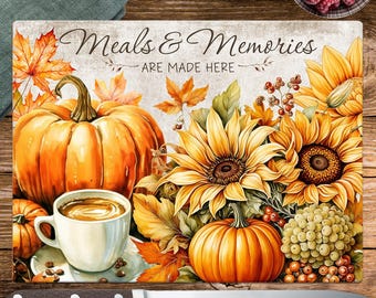May include: A decorative cutting board with a fall theme. It features a large orange pumpkin, sunflowers, and the text "Meals & Memories ARE MADE HERE." A white coffee cup and a knife are also visible.
