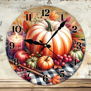 May include: Round wall clock featuring a pumpkin, gourds, apples, and berries. The clock face has black numerals and hands. A lit candle and fall leaves complete the autumn-themed design. The clock is set against a wood-grain background.