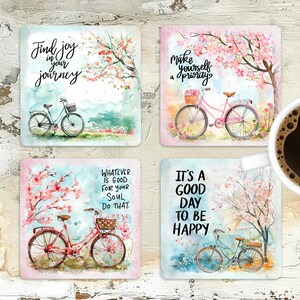 K&ouml;nnte beinhalten: Set aus vier quadratischen Untersetzern mit Aquarell-Blumenmustern und inspirierenden Zitaten. Jeder Untersetzer zeigt ein Fahrrad mit einem Blumenkorb und einen rosa-wei&szlig;en Kirschbl&uuml;tenbaum. Die Zitate lauten: "Finde Freude auf deiner Reise", "Mache dich selbst zur Priorit&auml;t", "Was immer gut f&uuml;r deine Seele ist, tu das", und "Es ist ein guter Tag, um gl&uuml;cklich zu sein".