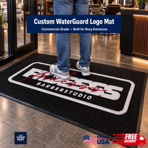 May include: A rectangular black floor mat with a textured surface and a white border. The center features the white text "FADEZ" in a bold font with a red script overlay, and the word "BARBERSTUDIO" in white capital letters below it.
