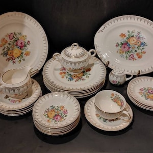 May include: A set of vintage floral china dinnerware, including a large platter, a serving bowl, a creamer, six saucers, six teacups, and six dessert plates. The floral pattern features pink, yellow, and white flowers on a white background.
