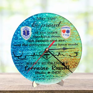 May include: A round glass clock with a blue, green, and yellow gradient background. The clock features a stethoscope, a heartbeat, and a medical symbol. The text "May you be proud of the work you have done, the person you are, and the difference you have made" is printed on the clock. The clock also features the text "Happy Retirement Lorraine Russell Medic # 0431 1/24/1982 - 4/1/2025".