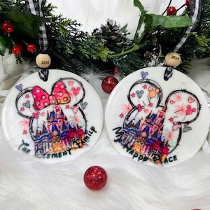 Pode incluir: Duas decorações de Natal redondas e brancas com um design de castelo da Disney. Uma apresenta um laço da Minnie Mouse, a outra orelhas do Mickey Mouse. Ambas têm o texto "The Family" e "My Happy Place". Cada enfeite tem uma fita preta e branca e uma conta de madeira com "2025".