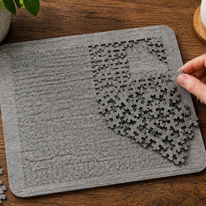 May include: A square gray wooden puzzle board with a textured surface featuring a grid of interlocking puzzle pieces. Some pieces are fitted into the board, while others are piled in the center and scattered near the bottom left corner.