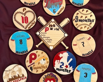 Może przedstawiać: Zestaw drewnianych znaczników kamieni milowych dla dzieci w tematyce baseballu. Każdy okrągły znacznik przedstawia wzór koszulki z imieniem i numerem gracza oraz słowem "miesięcy". Centralny znak w kształcie baseballu ma miejsce na dane dziecka.