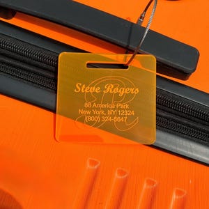 May include: Orange luggage tag with a clear plastic window. The tag has the name "Steve Rogers" and contact information: 88 America Park, New York, NY 12324, (800) 324-5647.