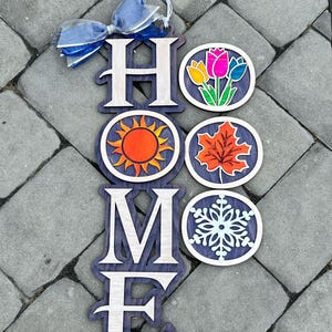 May include: A vertical wooden wall hanging spelling the word HOME with a blue ribbon at the top. The letter O is replaced by three interchangeable circular wooden plaques featuring a colorful tulip bouquet, an orange sun, a maple leaf, and a snowflake.