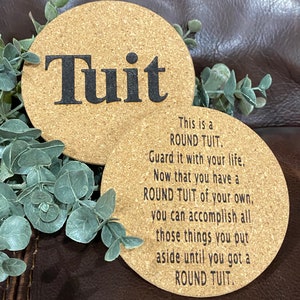 May include: Two round cork coasters with a saying about "round tuit". The top coaster has the word "Tuit" in large black letters. The bottom coaster has the text "This is a ROUND TUIT. Guard it with your life. Now that you have a ROUND TUIT of your own, you can accomplish all those things you put aside until you got a ROUND TUIT."