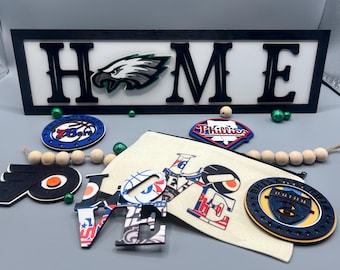 May include: A wooden sign with the word "HOME" in black letters on a white background. The sign is decorated with a Philadelphia Eagles logo, a Philadelphia Phillies logo, and a basketball logo. There are also several other decorative items, including a wooden "LOVE" sign, a fabric pouch, and a wooden bead garland.
