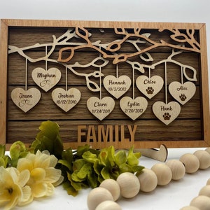 May include: A wooden family tree wall decor with hearts hanging from branches. Each heart has a name and date engraved on it. The names include Megan, Jason, Joshua, Hannah, Clara, Lydia, Chloe, Alex, and Hank. The text "FAMILY" is engraved at the bottom.