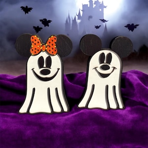Puede incluir: Dos decoraciones de Halloween de madera con forma de fantasmas con orejas de Mickey Mouse. Un fantasma tiene un lazo de lunares rojos y naranjas. El fondo presenta un castillo espeluznante, murciélagos y una superficie de terciopelo púrpura.