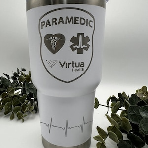 Może przedstawiać: Biały kubek termiczny ze stali nierdzewnej z grawerowanym wzorem w kolorze szarym. Wzór zawiera słowa "Paramedic", serce z symbolem medycznym, gwiazdę życia i słowa "Virtua Health". Na dole kubka znajduje się również szary grawerowany rytm serca.
