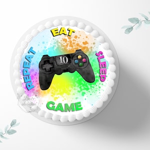 Könnte beinhalten: Ein runder Kuchen mit einer weißen Frosting-Basis und einem essbaren Bild in bunten Farben darauf. Das Bild zeigt einen schwarzen Videospiel-Controller mit einem Regenbogen-Spritzer-Hintergrund und dem Text "REPEAT EAT GAME SLEEP".