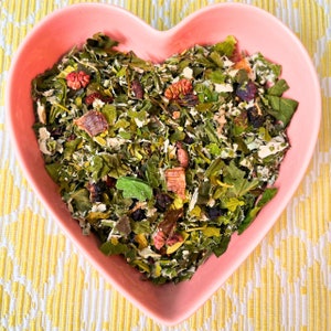 May include: A pink heart-shaped bowl filled with a blend of dried herbs, leaves, and berries. The bowl is sitting on a yellow and white patterned surface.