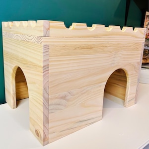 May include: A light-colored wooden castle-shaped structure with arched openings and a crenelated top. The wood grain is visible, and the structure appears to be handmade. The design is reminiscent of a medieval castle.