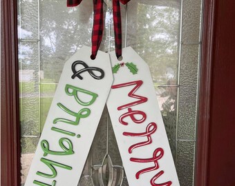 May include: Two white wooden signs with red and green lettering hanging from a red and black checkered bow. The signs read "Merry" and "Bright &"