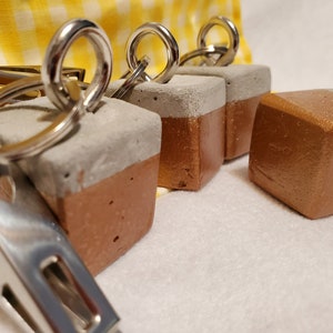 May include: Four concrete keychains with a two-tone color scheme. Each keychain is a cube with a gray top and a copper bottom. The keychains are attached to silver key rings.