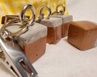 May include: Four concrete keychains with a two-tone color scheme. Each keychain is a cube with a gray top and a copper bottom. The keychains are attached to silver key rings.