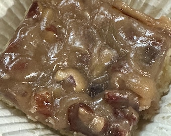 May include: A square pastry with a glossy, light brown topping, studded with nuts and served in a white paper cup. The pastry has a golden-brown crust and a variety of nuts, including pecans and cashews.