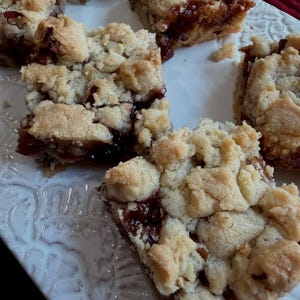May include: Several square-shaped fruit crumble bars arranged on a white plate. Each bar features a golden-brown, crumbly streusel topping with visible pieces of nuts, layered over a dark, jam-like fruit filling that is visible between the crumbs.