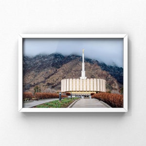 Peut inclure: Photographie encadrée d'un temple avec une haute flèche surmontée d'une statue dorée. Le temple a une conception circulaire avec des panneaux en bois verticaux. Des montagnes et un ciel nuageux forment la toile de fond. L'image est encadrée dans un cadre blanc.