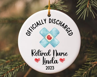 May include: White ceramic Christmas ornament with the text "Officially Discharged Retired Nurse Linda 2023" and a blue bandage with a red heart in the center.