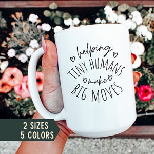 May include: White ceramic mug with black text that reads "helping tiny humans make big moves" with small hearts around the text. The mug is being held by a hand with red nail polish. The background is a blurry image of a flower garden.