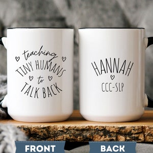 May include: Two white ceramic mugs with black handles and rims. One mug reads "teaching TINY HUMANS to TALK BACK" in black script. The other mug says "HANNAH" and "CCC-SLP". Both mugs have a heart symbol.