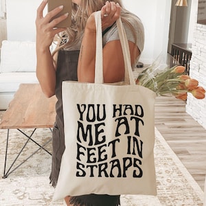 Puede incluir: Bolso tote color crema con el texto negro "YOU HAD ME AT FEET IN STRAPS". El bolso tiene asas largas y lo sostiene una persona. El fondo muestra un sal&oacute;n con un sof&aacute; y una chimenea.