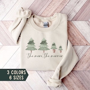 Puede incluir: Una sudadera beige con un diseño de acuarela de cuatro árboles de Navidad verdes con nieve. Los árboles están en fila, disminuyendo de tamaño de izquierda a derecha. El texto "the more, the merrier" está debajo de los árboles.