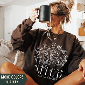 May include: A brown sweatshirt with a white floral design and the text "girls gone mild asheville 2025".