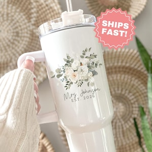 Puede incluir: Vaso blanco con asa, dise&ntilde;o floral y el texto "Mrs. Johnson EST. 2026". El vaso tiene una tapa transparente y una pajita. El dise&ntilde;o incluye flores blancas y verde claro. El vaso lo sostiene una persona que lleva un su&eacute;ter blanco.