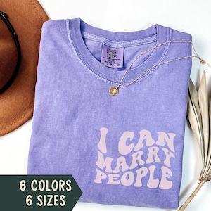 Puede incluir: Camiseta morada con el texto "I can marry people" en letras rosas onduladas. La camiseta está hecha de un material suave y cómodo y está disponible en 6 colores y 6 tallas.