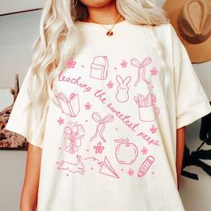 May include: Ivory-colored t-shirt with a pink graphic design. The design includes a backpack, book, flowers, bows, a bunny, a pencil, an apple, and the text "Teaching the sweetest peeps."