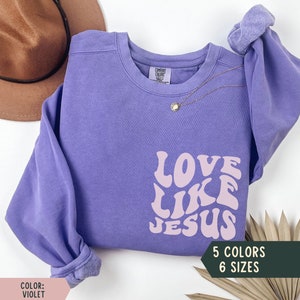K&ouml;nnte beinhalten: Lilafarbener Pullover mit dem Text "LOVE LIKE JESUS" in rosafarbenen Retro-Lettern. Der Pullover ist in 5 Farben und 6 Gr&ouml;&szlig;en erh&auml;ltlich.