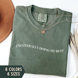 K&ouml;nnte beinhalten: Ein olivgr&uuml;nes T-Shirt mit dem wei&szlig;en Aufdruck "I'M LITERALLY DOING MY BEST". Eine goldene Halskette mit Anh&auml;nger liegt auf dem Shirt. Das Bild zeigt auch einen braunen Hut und Text, der sechs Farben und sechs Gr&ouml;&szlig;en angibt.