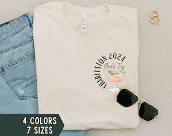 Könnte beinhalten: Ein weißes T-Shirt mit einem schwarzen Kreisdesign, auf dem "Charleston 2024 Girls Trip" steht und ein rosafarbener Cocktail im Kreis ist. Das T-Shirt liegt auf einer beigefarbenen Oberfläche mit einer blauen Jeans und einem Paar goldenen Creolen.
