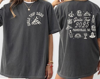 Op de afbeelding: Donkergrijs t-shirt met witte grafische tekst en illustraties. De voorkant van het shirt zegt "Girls Trip 2025" boven een berg- en maanontwerp. De achterkant van het shirt heeft illustraties en de tekst "Girls Trip 2025 Asheville, NC".