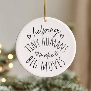 May include: A round white ceramic ornament hanging from a gold string. The ornament features black text that reads: helping TINY HUMANS make BIG MOVES, accented with four small black heart outlines positioned around the central lettering.
