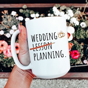 May include: White ceramic coffee mug with black text that reads "Wedding Lesson Planning." A pencil is drawn through the word "Lesson" and two gold rings are drawn above the word "Wedding".