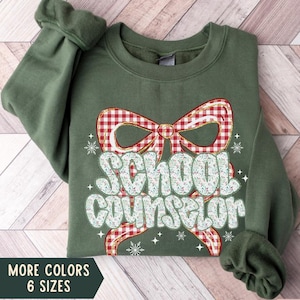 Puede incluir: Sudadera verde militar con la frase "School Counselor" en una fuente lúdica, adornada con un lazo a cuadros rojo y blanco y copos de nieve. El diseño incluye "MORE COLORS 6 SIZES".