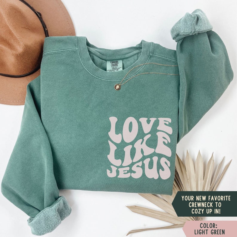 K&ouml;nnte beinhalten: Ein hellgr&uuml;ner Pullover mit den Worten "LOVE LIKE JESUS" in einer rosafarbenen Retro-Schrift.