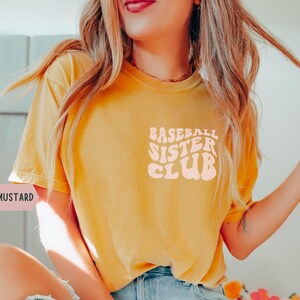 May include: Mustard-colored t-shirt with the text "Baseball Sister Club" in a retro font. The shirt is a solid color and has short sleeves. The text is in a light pink color.