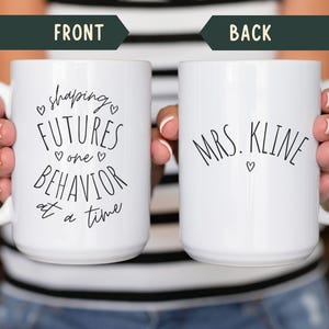 May include: White ceramic mug with black text that reads "shaping FUTURES one BEHAVIOR at a time" on the front and "MRS. KLINE" on the back. The mug has a white handle and is held by a person.